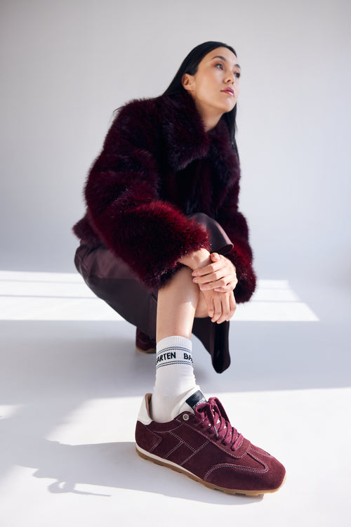 Person wearing a dark fur coat and maroon sneakers sitting on a white surface.