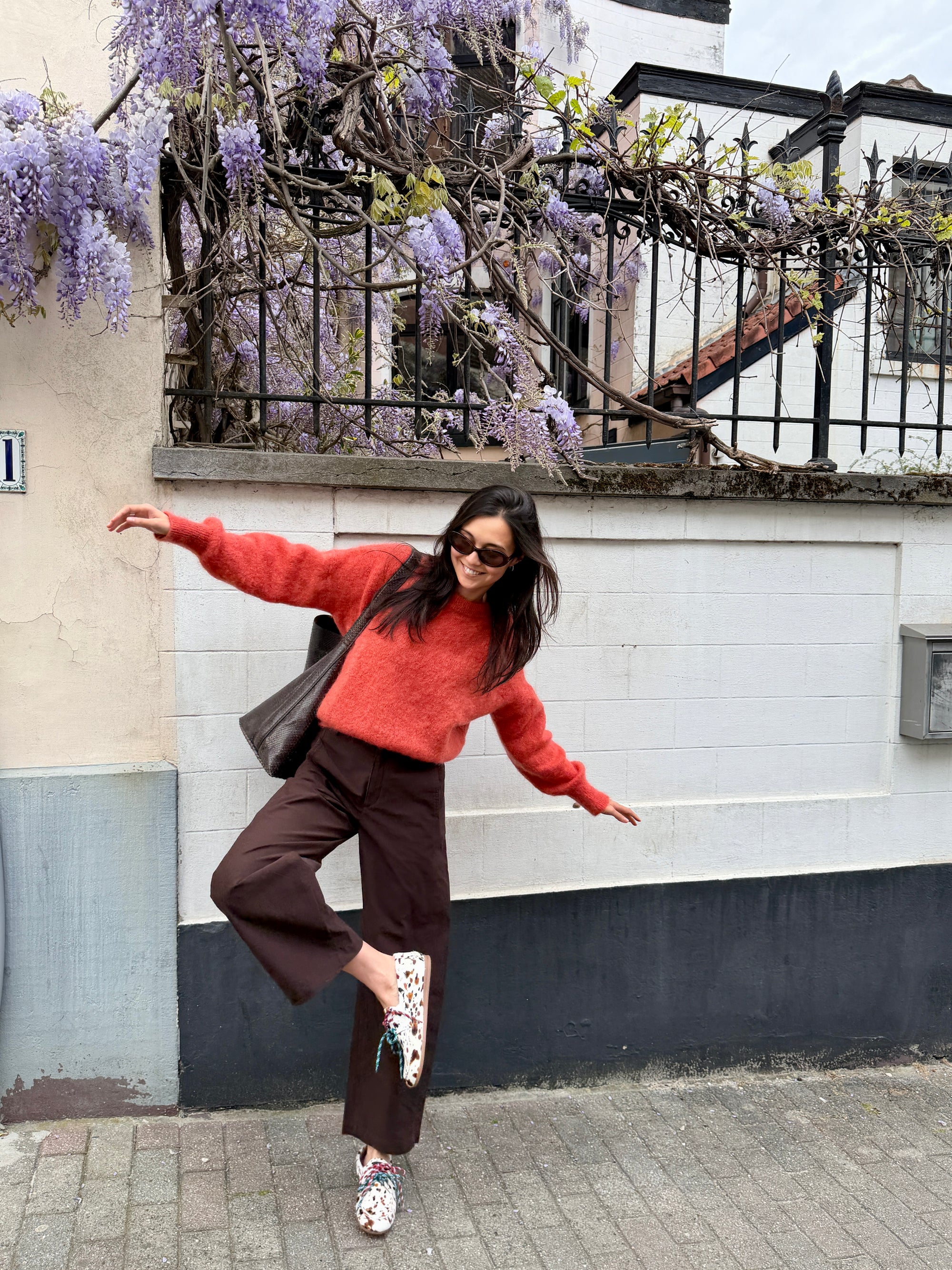 Woman in a red sweater and brown pants dancing in front of a building with wisteria